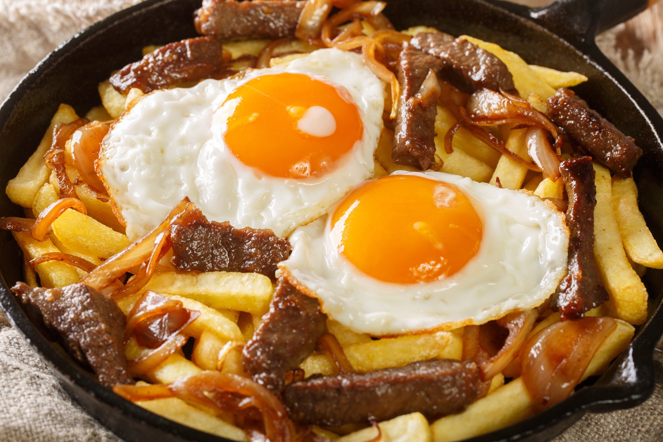 Chorrillana is a dish consisting of french fries topped with sliced meat, fried eggs, and onions close up in the pan. Horizontal